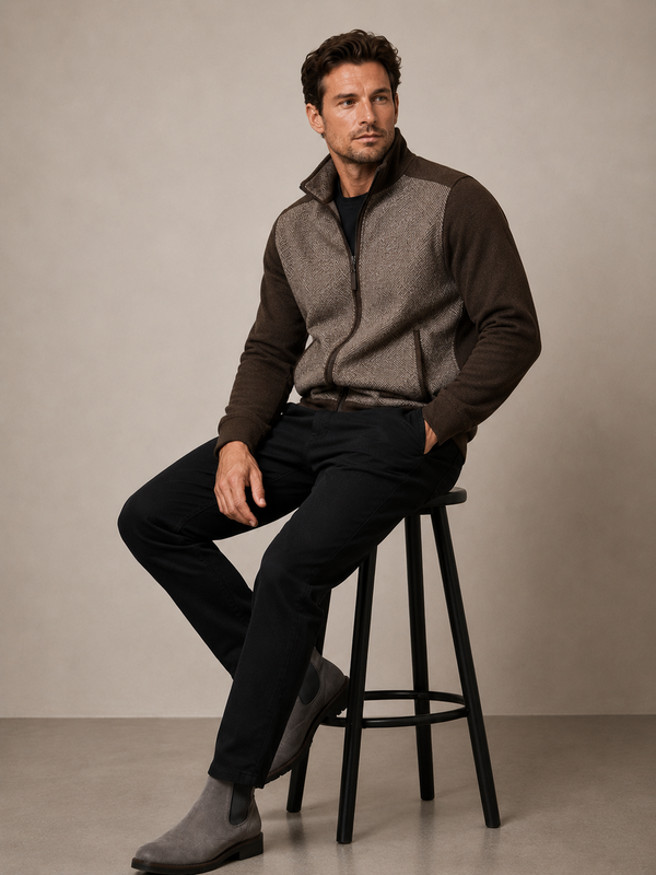 A man sits on a black stool wearing a brown and gray quarter-zip fleece jacket, black trousers, and gray suede boots.